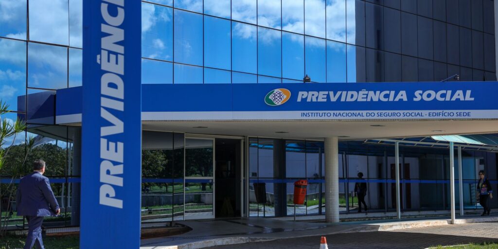 AGU cobra R$ 135 milhões em último lote de ações contra gigantesca fraude no INSS 2 Vista aérea de um moderno edifício de escritórios em um centro urbano, apresentando uma fachada de vidro reflexiva com múltiplos andares e geometria angular. O céu está parcialmente nublado, e a iluminação lateral destaca a estrutura, sugerindo um ambiente formal e institucional, como o de órgãos governamentais ou entidades jurídicas.